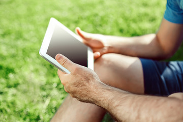 Close-up of casual dressed young man using modern digital tablet while sitting at the park, sun light