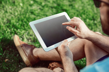 Close-up of casual dressed young man using modern digital tablet while sitting at the park, sun light