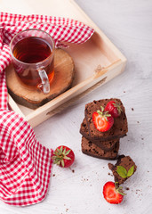Hot black tea, delicious homemade chocolate brownie pieces decorated with organic strawberries on wooden box and concrete table. Top view, copy space, close up.