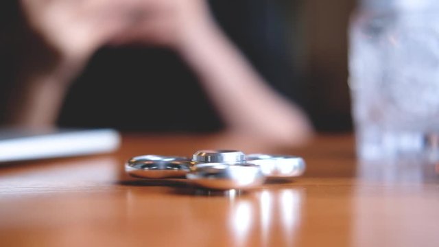 A metal silver color fidget spinner is spinning on wooden table with a person in background