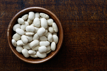 Fototapeta na wymiar Dry butter beans in dark wooden bowl isolated on dark brown wood from above.