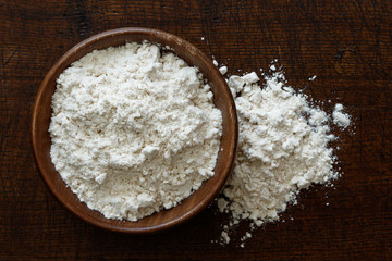 Fototapeta na wymiar Fine white flour in dark wooden bowl isolated on dark brown wood from above. Spilled flour.