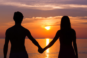 Romantic Valentine's Day scene of a young couple silhouettes holding hands by the sea staring at...