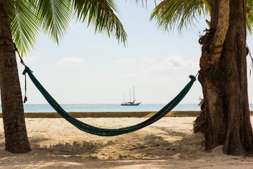 Beach hammock hanging from a thick tree and a palm tree with two sailboats on the background in the island of Koh Phangan, Thailand.