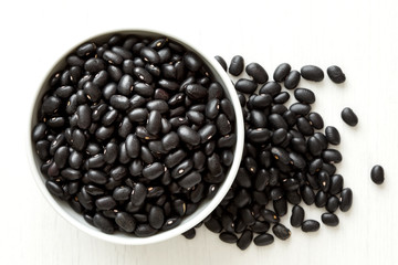 Black beans in white ceranic bowl isolated on painted white wood from above. Spilled beans.