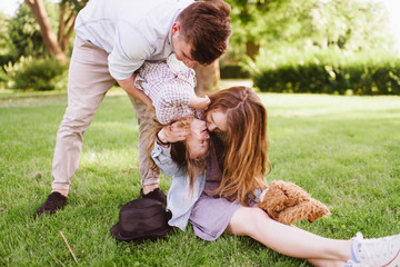 Fototapeta premium Young cheerful family sitting on grass enjoying vocation and playing with their child, father holding daughter upside down. People sitting in summer park. Sunny day, outdoor