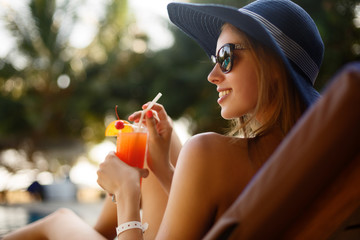 Portrait of young woman with cocktail glass chilling in the tropical sun near swimming pool on a...