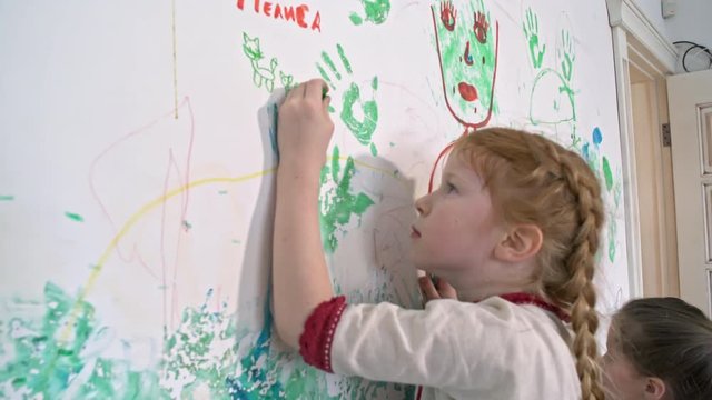 Side view of little redhead girl with braided hair and her older sister doodling with crayons on wall covered in drawings