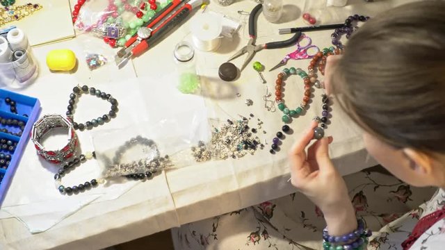 PAN with top view of young woman sitting before table with tools and making bracelet from beads
