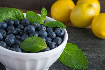 Closeup of fresh, sweet blueberries in a stylish white bowl with green mint herb leaf and yellow lemon in the background, placed on a wooden background