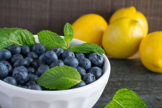 Delicious summer fruit - Fresh, Sweet blueberries in a stylish white bowl with mint herb leaf and lemon in the background