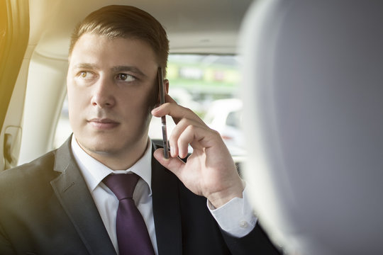 Businessman working while sitting in a car. A man is driven to work in his limo. Suit and tie businessman in the back seat making a call while the limo driver is driving. Lens flare in the background.