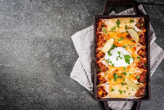 Mexican food. Cuisine of South America. Traditional dish of spicy beef enchiladas with corn, beans, tomato. On a baking tray, on a black stone background. Top view copy space