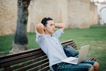 Young asian man sitting on the park bench with laptop and relax with good mood after finished work