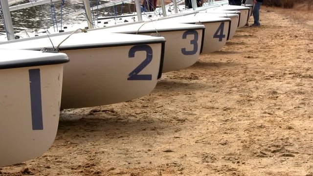A line of 420 racing sailboats wait on shore of lake for rigging by high school sailing team.  Slight zoom-in along front of boats