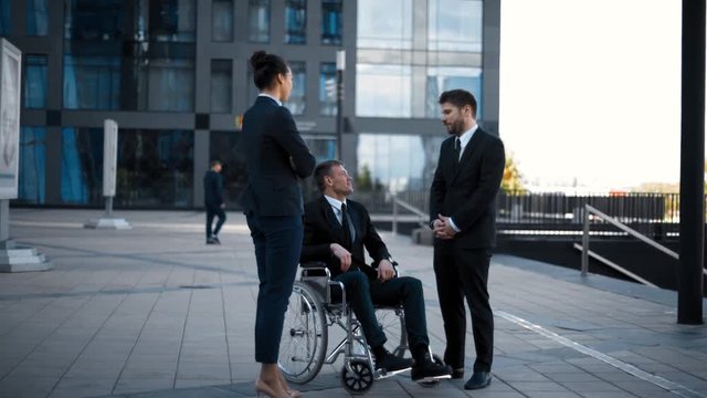 Handicapped invalid caucasian man in wheelchair and his business collegues has conversation near office modern building at sunset.