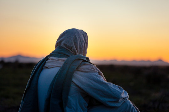 Jesus Christ sits on a hill side and watches the sun set on the horizon.