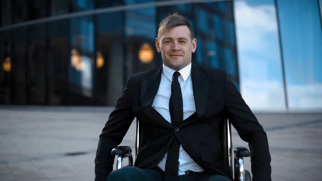 Disabled attractive invalid entrepreneur unfolds on wheelchair outdoor. Armchair on wheels. Handicapped man near glass building offices. Happy emotion portrait face middle shot