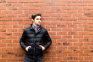 Cool fashion man sitting with his hand in pocket smiling and looking against brick wall