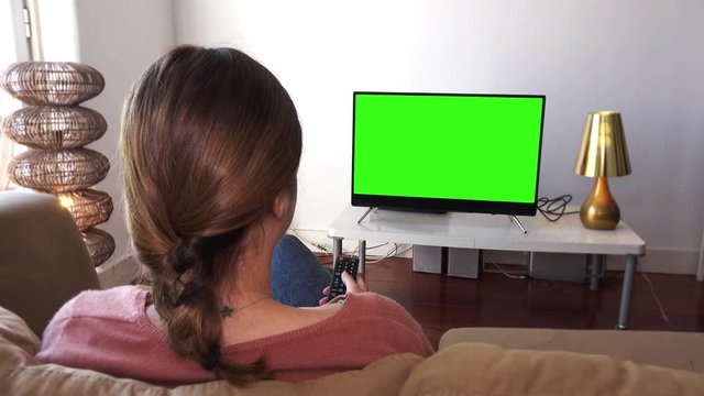 Woman Using Television Remote In Green Screen. Young Woman watching television with green screen, shot behind models shoulders