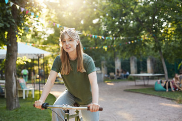 Portrait of young beautiful blonde woman enjoying pretending to ride a bicycle in the park during a food festival smiling on camera