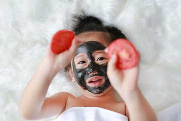 Kid girl in coal peeling face mask holding tomato slices on hands, beauty concept.