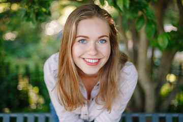 Closeup portrait of beautiful smiling white Caucasian girl woman with long blonde  hair and blue eyes, wearing white shirt and jeans, outside in summer park in green foliage trees, looking in camera.