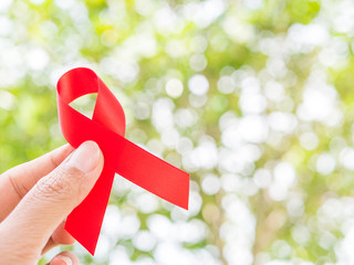 World AIDS day concept. Hand holding red ribbon on tree leaves bokeh background.