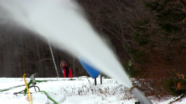 A medium shot of snow making gun at Bretton Woods in white mountain region of New Hampshire.  Snow rains down on ski trail as skiers can be seen in the background along edge of trail.  Includes audio