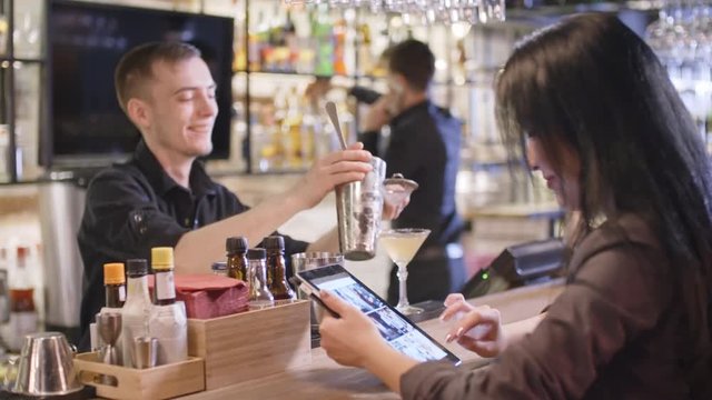 PAN of cheerful male bartender pouring cocktail from shaker tins into martini glass and chatting with smiling Asian woman sitting at counter and looking at pictures on tablet 