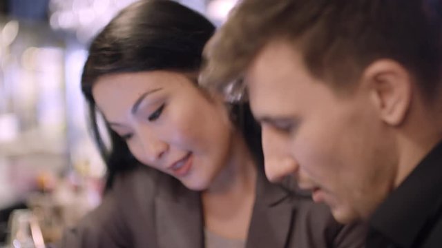 Tilt down of beautiful Asian businesswoman sitting at bar counter and discussing pictures on tablet with young male business partner 