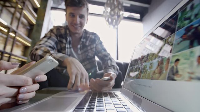 Young man showing pitching new project and showing pictures on laptop with unrecognizable woman with mobile phone during meeting in casual atmosphere 