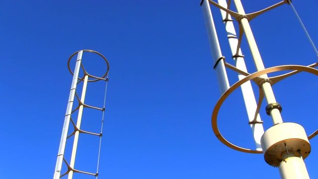 A pair of vertical axis wind turbines utilize renewable wind energy while spinning against blue sky.  These virtually silent windspires are made from recycled materials and stand 30' high