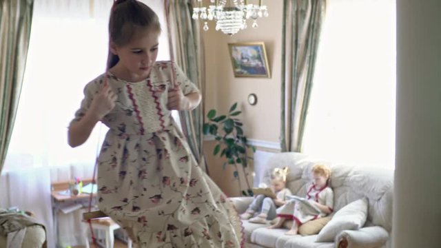 Tilt up of little girl in linen dress swinging on trapeze hanging from ceiling of living room; her siblings sitting on sofa and playing on tablets in background