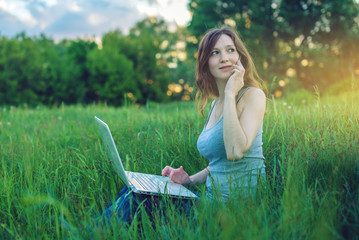 Woman sitting on a green meadow on the background of sunset with clouds. Working on laptop and talking on the phone.