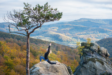 Sporty fit girl is practicing yoga and doing asana Virabhadrasana 1 on the top of the mountain near big tree. Autumn forests, rocks and hills on the background