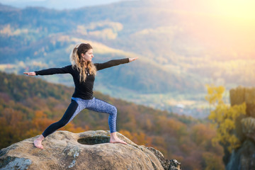 Attractive slim girl is practicing yoga and doing asana Virabhadrasana 2 on the top of the mountain in the evening. Autumn forests, rocks and hills on the background