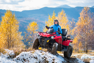 Smiling woman in blue jacket riding a red quadbike on snow-covered hill at sunny day against trees with yellow leaves and mountains. Blurred background