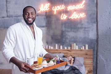 Delightful African American man holding breakfast tray in the bedroom