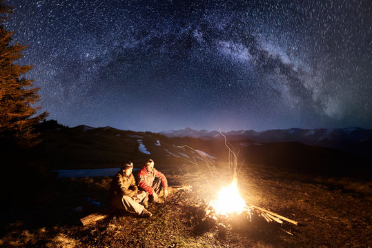 Two male hikers have a rest in the camping at night. Men sitting near campfire and tent under beautiful night sky full of stars and milky way, and enjoying night scene. Long exposure