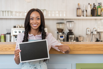 afro-american waitress pointing at empty board and looking at camera while standing at cafe