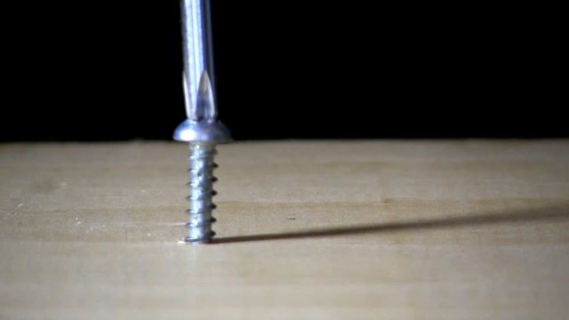 A pan head screw is tighten in pine board by hand with phillips head screwdriver on left side.  Macro shot from slight high angle looking down with black background.