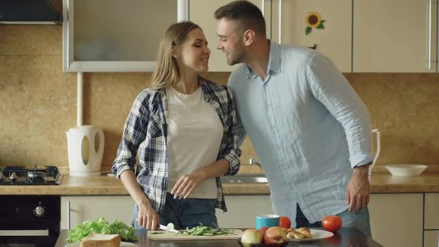 Happy young couple in the kitchen. Attractive woman cooking breakfast and cuff her boyfriend who want to taste food