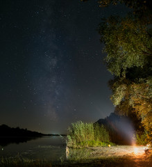 Fototapeta na wymiar The milky way over the water of the river, the fisherman sitting on the beach with bonfire