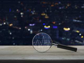 Magnifying glass with graph on wooden table over blur colorful night light city tower background,...