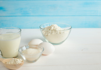 Baking in the kitchen - the ingredients of the test recipe (eggs, flour, milk, sugar) on a white wooden table with the inscription on top. Background layout with free text space.