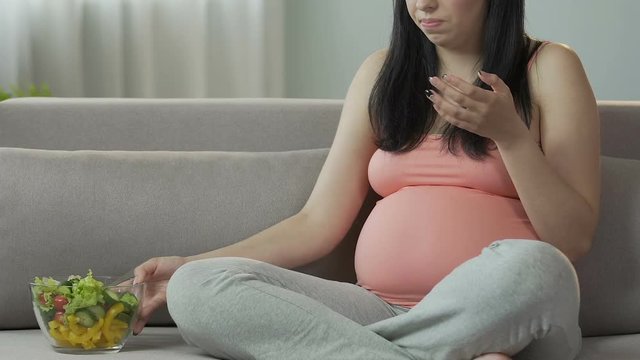 Expecting woman eating salad on couch and having fit of nausea, food sickness