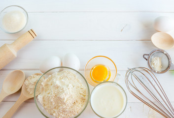 Baking in the kitchen - the ingredients of the test recipe (eggs, flour, milk, sugar) on a white wooden table with the inscription on top. Background layout with free text space.