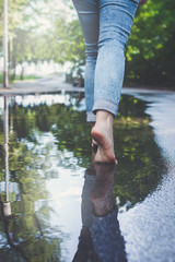 Barefoot woman in blue jeans walking through puddle of water in urban setting surrounded by trees 
