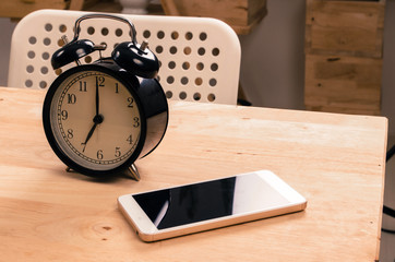 Naklejka premium smartphone and black retro alarm clock times at 7 o'clock morning on wooden table, selective focus, copy space, vintage color tone